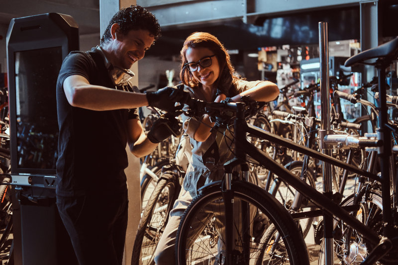 Viele Fahrräder mit einem Geschäft in Hintergrund. Ein Mann und eine Frau schrauben am Lenker des Fahrrads, welches in einer Montage hängt.