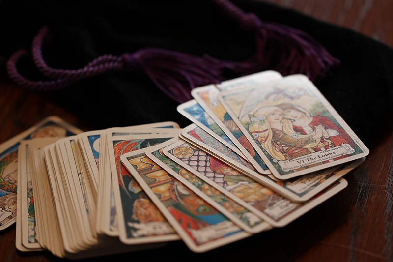 Ein Deck aus Tarotkarten ist auf einem Tisch längs ausgebreitet worden. Die Karte "Die Liebenden" ist an der Spitze zu erkennen. Der Hintergrund ist verschwommen und zeigt eine violette Kordel.